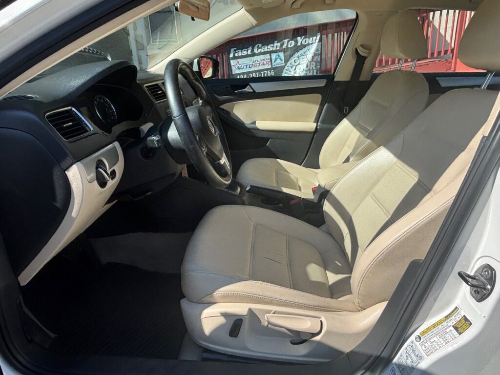 Beige interior of a sedan: driver and front passenger seats, steering wheel, and dashboard visible from the open driver’s door in daylight.