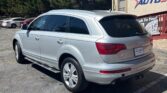 Rear three-quarter view of a silver Audi SUV parked in a car dealership lot, with a beige building and sign in the background.
