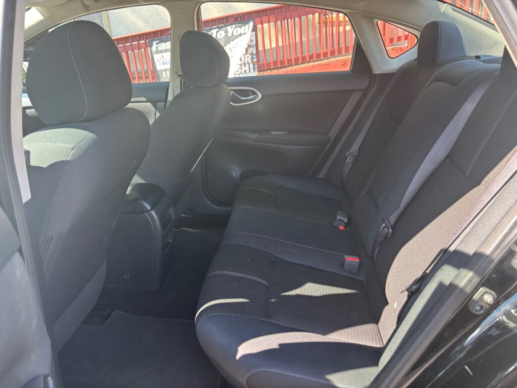 Interior of a dark fabric-sedan back seat area with a three-seat bench and visible seat belts, front seat in view, Sunlit window on the right.