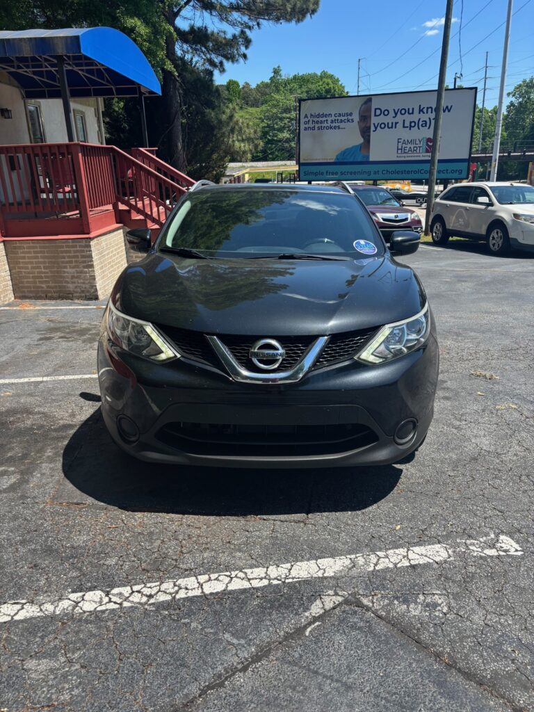 Front view of a black Nissan SUV parked in a lot with a blue awning-covered building to the left and a billboard in the background.