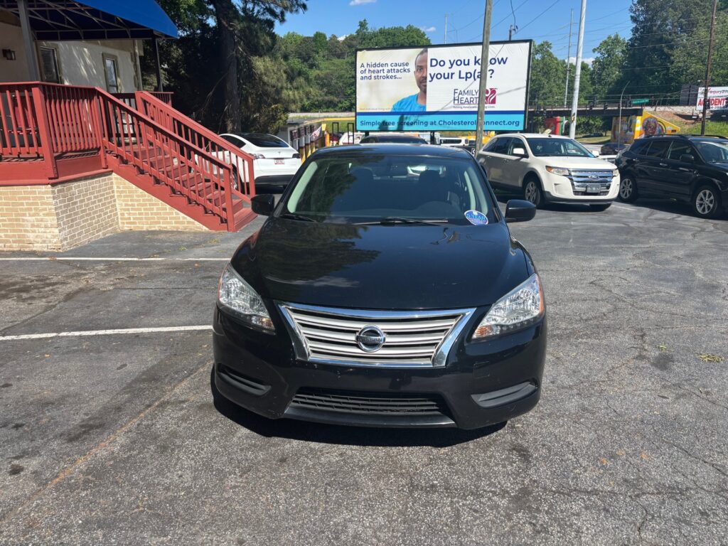 Front view of a black Nissan sedan parked in a lot beside a brick building and a large blue billboard in the background.