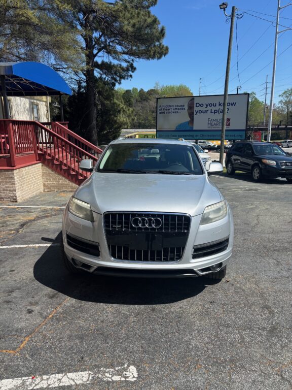 Front view of a silver Audi SUV parked in a lot beside a brick building with a blue canopy and red railing; large billboard in the background.
