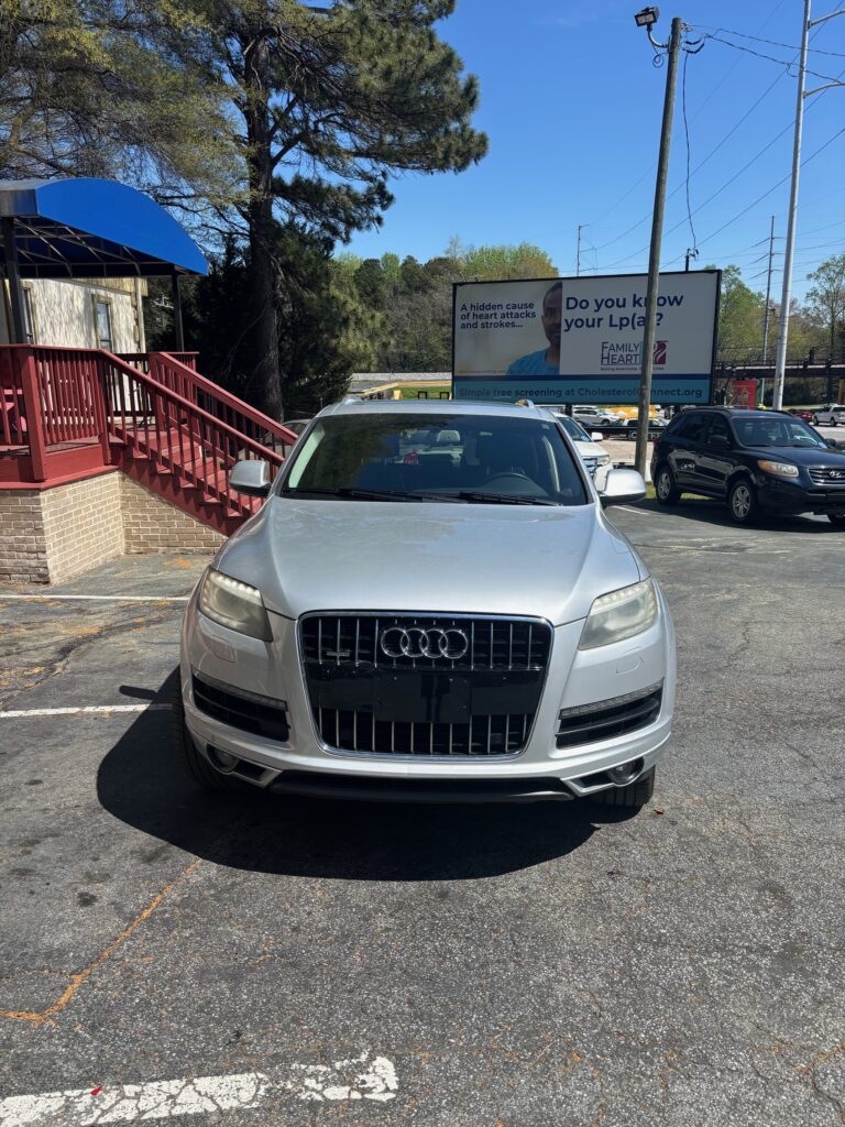 Front view of a silver Audi SUV parked in a lot beside a brick building with a blue canopy and red railing; large billboard in the background.