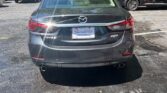 Rear view of a gray Mazda sedan parked in a sunny parking lot with other cars nearby.