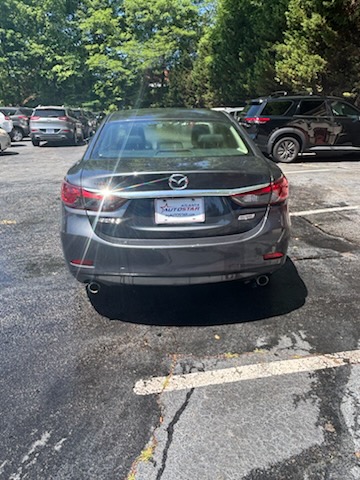 Rear view of a gray Mazda sedan parked in a sunny parking lot with other cars nearby.