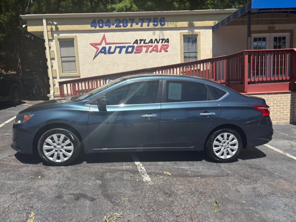 Side view of a blue-gray sedan parked in front of the Atlanta AutoStar dealership building with a red railing and beige exterior.