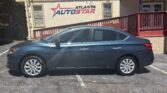 Side view of a blue-gray sedan parked in front of the Atlanta AutoStar dealership building with a red railing and beige exterior.
