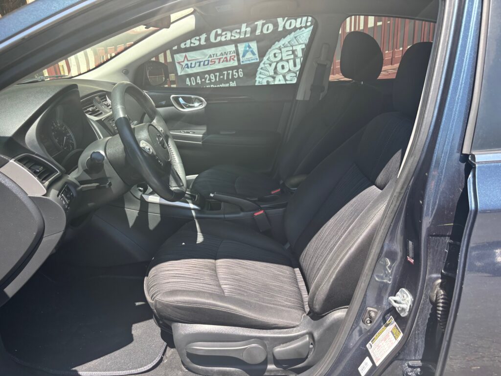 Interior view of a dark blue sedan from the open driver's side door, showing black cloth front seats, steering wheel, dashboard, and center console.