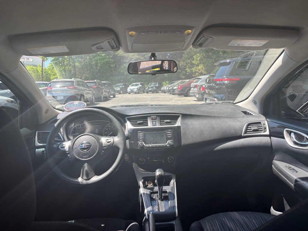Interior of a car viewed from the back seat, showing the steering wheel, center console, and dashboard with a parking lot of cars outside through the windshield.