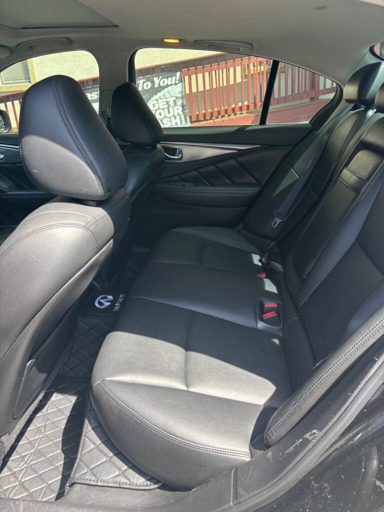 Rear passenger area of a black leather sedan, with a dark bench seat, three headrests, and quilted floor mats visible in sunlight.