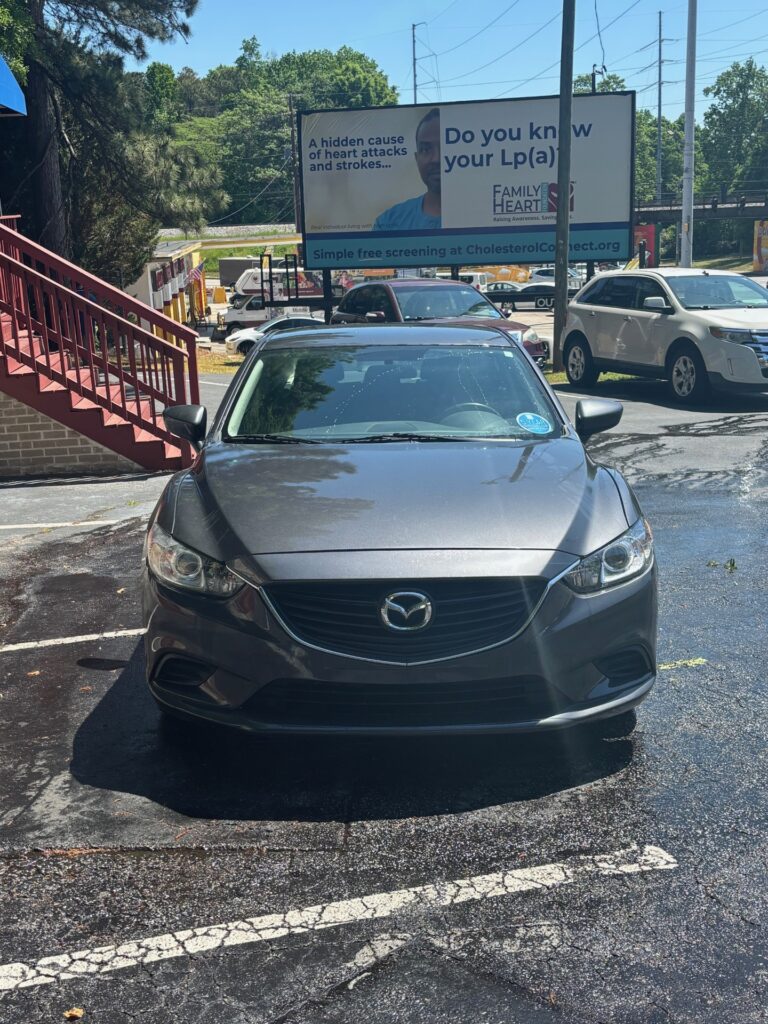 Front view of a gray Mazda sedan parked in a lot with a red staircase on the left and a large billboard in the background advertising cholesterol screening.