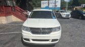 Front view of a white minivan parked in a lot, with a brick building and blue awning to the left and a billboard in the background.