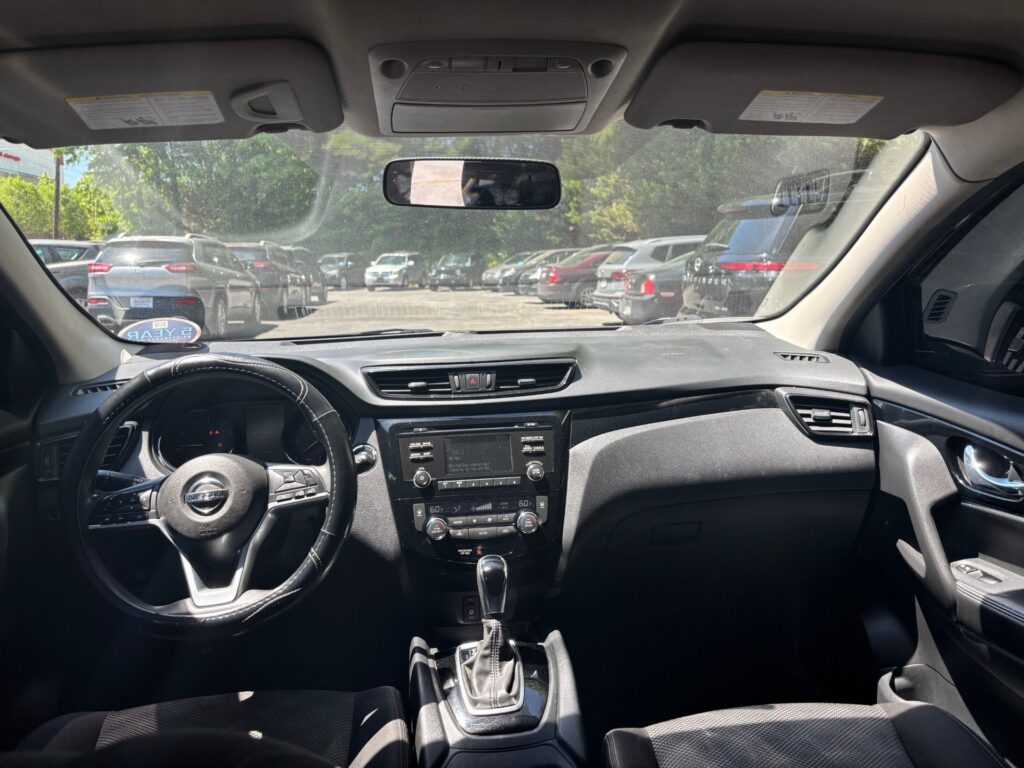 Interior view from the driver’s seat: steering wheel, center console with radio, and gear shifter, parking lot seen through the windshield with multiple cars