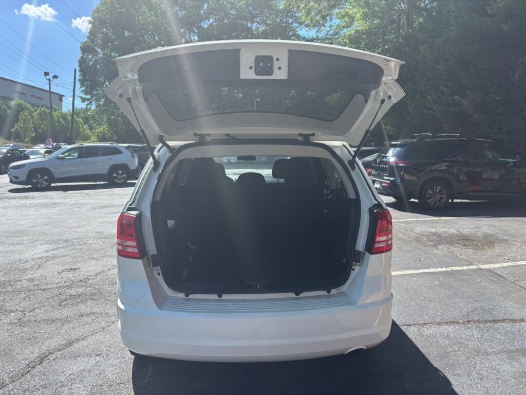 White SUV with its tailgate open, showing the dark cargo area in a sunny parking lot. Gobetween vehicles and trees in the background.
