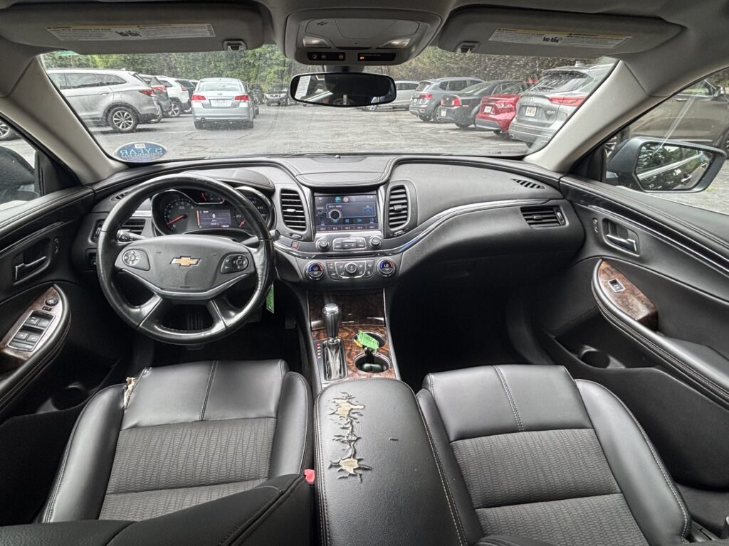 Front car cabin with steering wheel, dashboard touchscreen, and center console; black leather seats with a torn armrest cover in the foreground.