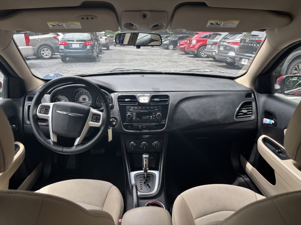 View from the back seat of a beige-car interior showing steering wheel, center console, and gear shifter facing a parking lot through the windshield.
