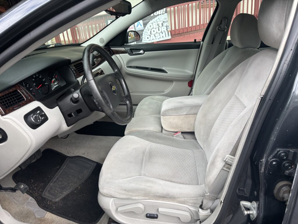 Interior of a gray fabric car cabin: steering wheel with Chevy logo, dashboard, and front seats; view from the open driver’s side door with a window poster outside.