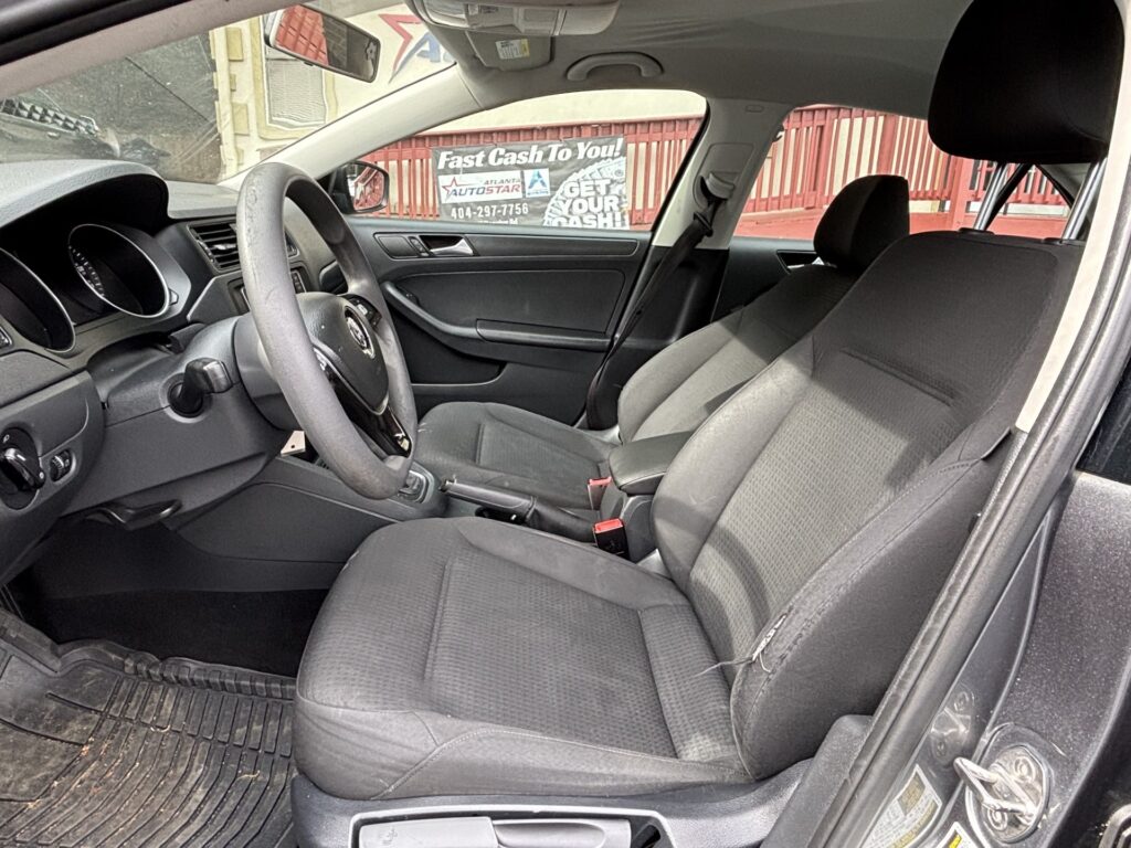 Interior of a compact car showing the driver's seat, steering wheel, and front passenger seat with dark upholstery and floor mats. The view is from the open passenger side door.
