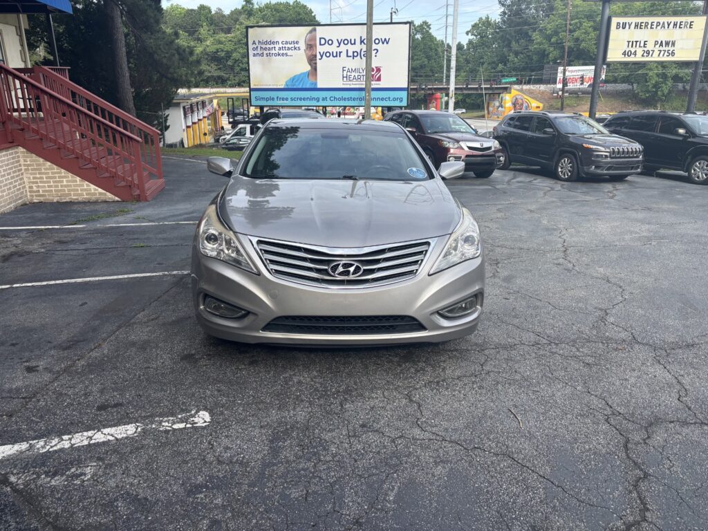 Front view of a silver Hyundai in a parking lot with other cars and a red staircase on the left. Nearby billboards and trees in the background.