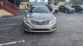 Front view of a silver Hyundai in a parking lot with other cars and a red staircase on the left. Nearby billboards and trees in the background.