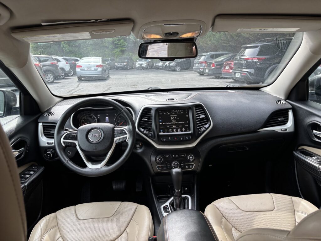 Interior view of a Jeep SUV showing the steering wheel, center console touchscreen, and beige leather seats from the back seat. rearview mirror reflections visible, sun visors up.