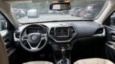 Interior view of a Jeep SUV showing the steering wheel, center console touchscreen, and beige leather seats from the back seat. rearview mirror reflections visible, sun visors up.