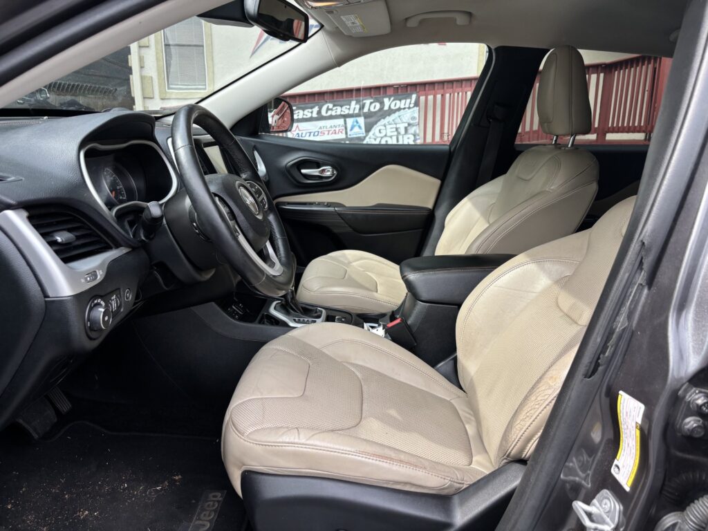 Front interior of a Jeep with beige leather seats, steering wheel, and center console; view through the passenger door shows a red fence and a 'Fast Cash To You' banner outside.