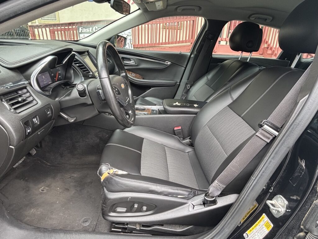 Front passenger and driver seats of a black leather car interior; visible wear on the driver seat with torn upholstery and exposed foam. Steering wheel, dashboard, and center console in view.