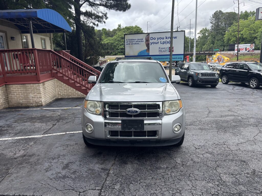 Front view of a silver Ford Escape parked in a lot, with a brick building and blue awning on the left and a large billboard in the background.