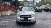Front view of a silver Ford Escape parked in a lot, with a brick building and blue awning on the left and a large billboard in the background.