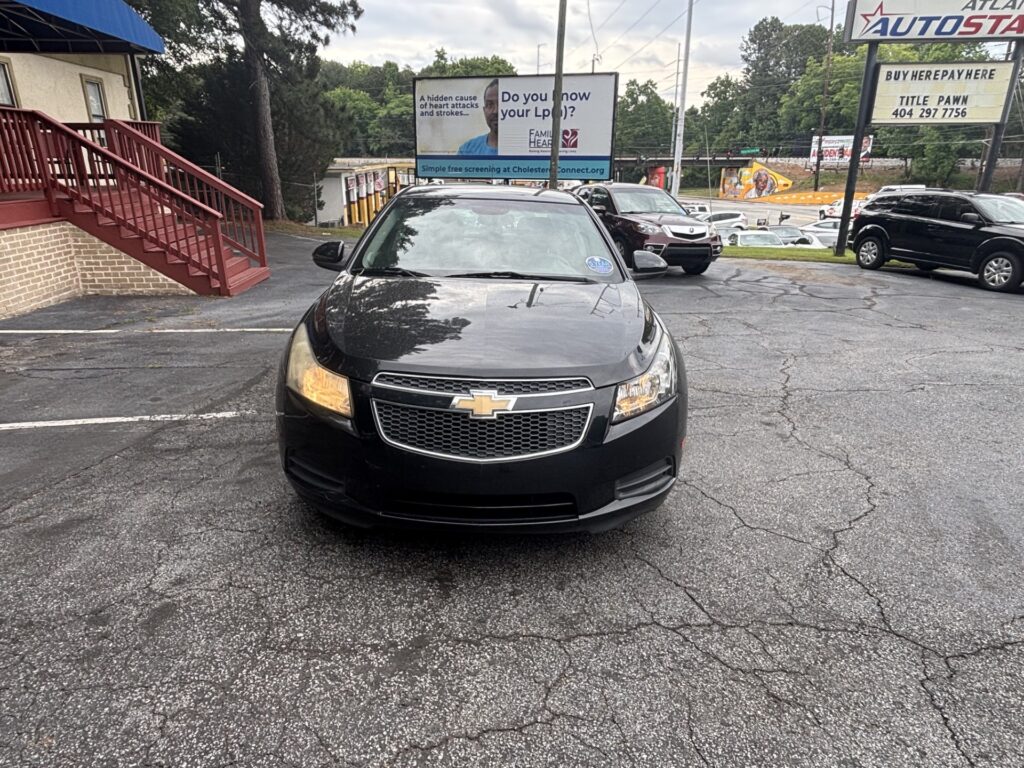 Front view of a black Chevrolet vehicle centered in a parking lot, with a brick building and red stairs on the left and billboards in the background.