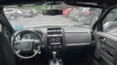 Front interior of a car showing left-hand steering wheel, center touchscreen, gear shift, and black leather seats while parked in a lot.
