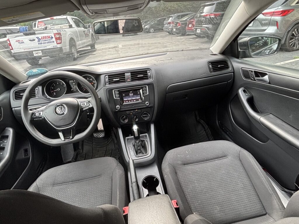 Interior of a car from the front passenger seat: steering wheel, dashboard, center console, and gear lever visible with fabric seats and cup holders between them. Outside, parked cars are seen through the windshield and side windows.