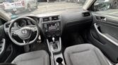 Interior of a car from the front passenger seat: steering wheel, dashboard, center console, and gear lever visible with fabric seats and cup holders between them. Outside, parked cars are seen through the windshield and side windows.