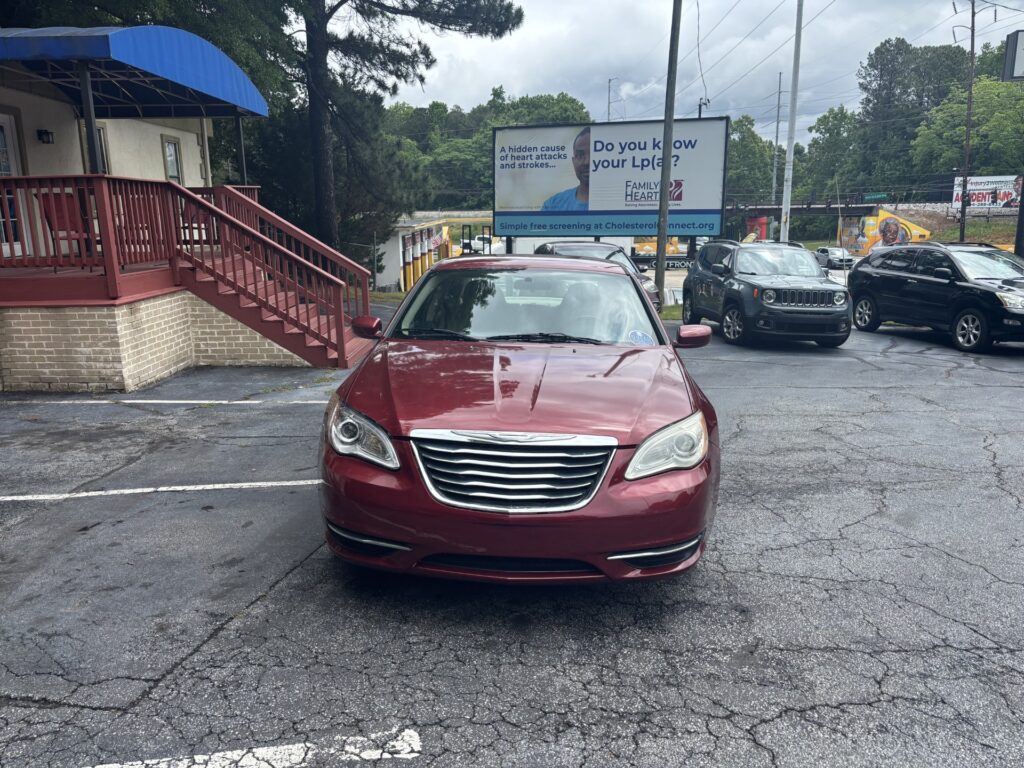 Red sedan parked head-on in a parking lot, with a brick building and blue awning on the left and a large billboard in the background promoting cholesterol screening.
