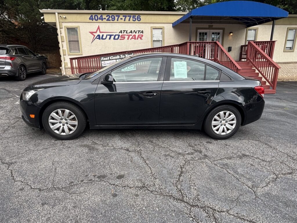 Black sedan parked in a dealership lot in front of a beige building with a blue awning and red stairs, window signs visible on the car's side window.