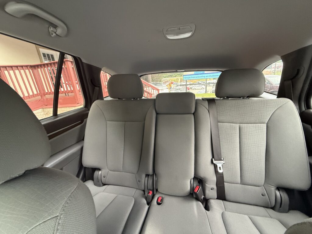 Interior view of a car's rear row with three grey seats and visible seat belts; outside shows a red railing and parking area.