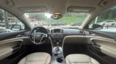 Interior of a beige car cabin viewed from the back seat, showing the dashboard, steering wheel, and center console. Outside windows reveal a parking lot with other cars and greenery.
