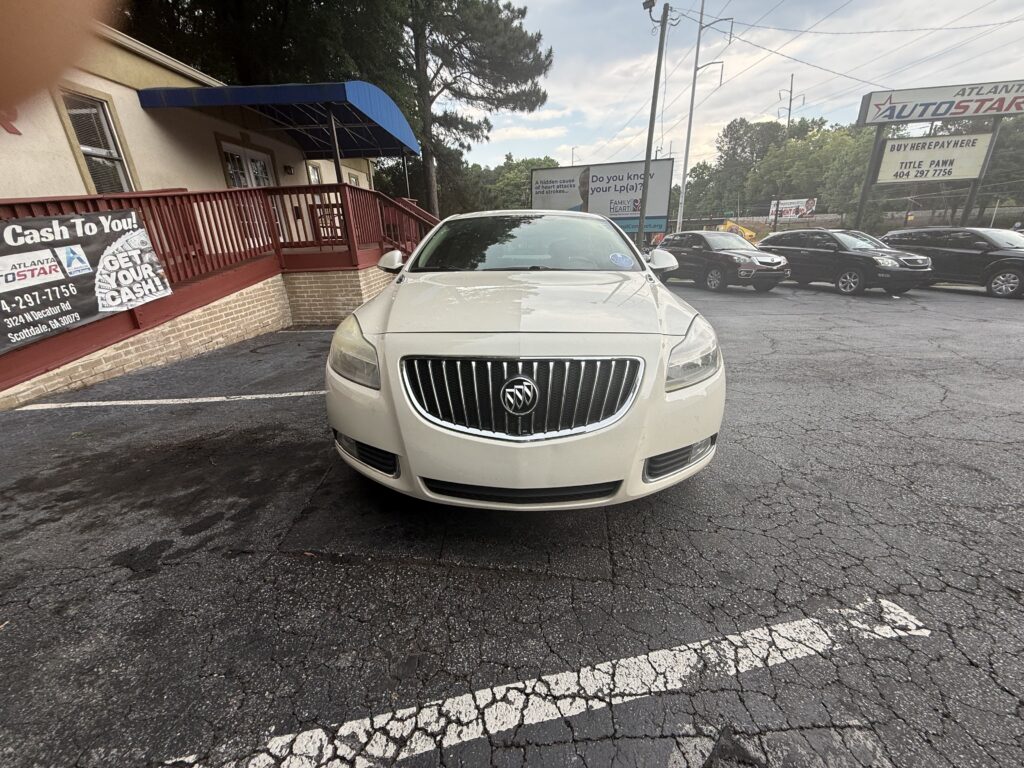 White Buick SUV front and center in a dealership lot, with a brick building and blue awning to the left and advertising banners along the railing.