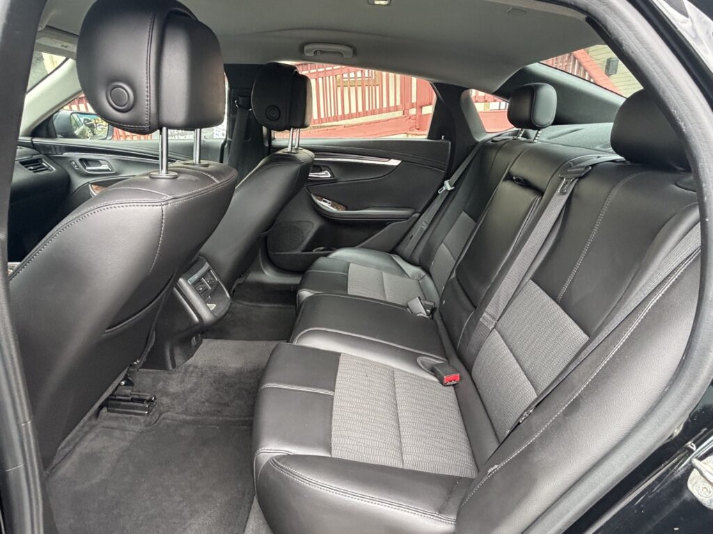 Rear passenger seating area of a black leather car interior with two headrests and visible seatbelts. The door panel and wood trim are also shown.