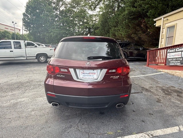 Rear view of a maroon Acura MDX SUV parked in a lot, showing the dealership plate frame and surrounding cars in the background.