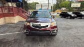 Maroon Acura SUV photographed from the front in a car dealership lot, with signs and other vehicles visible in the background.