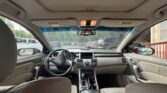 Interior of a beige sedan viewed from the back seat, showing the steering wheel, center console, and front windshield with outside traffic seen through it.