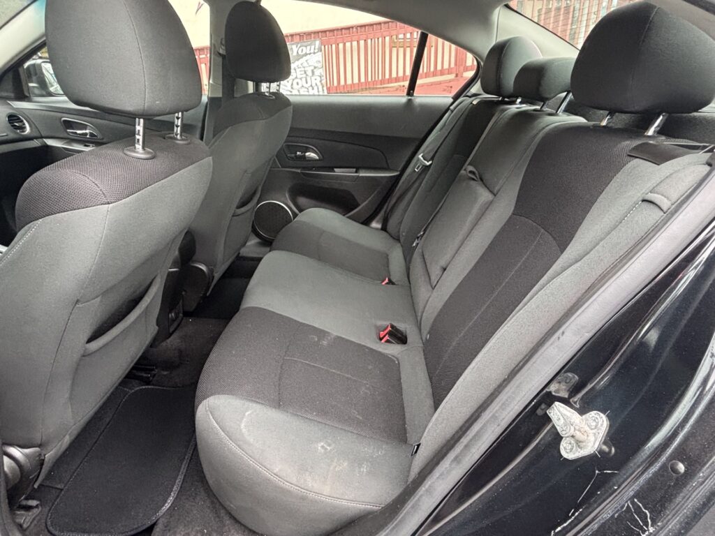 Rear interior of a dark fabric sedan: back seats with headrests, seat belts, and door panel visible through an open door edge.