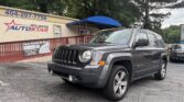 Gray Jeep Patriot parked in front of Atlanta AutoStar dealership under a blue awning. The building displays the 'Atlanta AutoStar' sign and posters in the foreground.