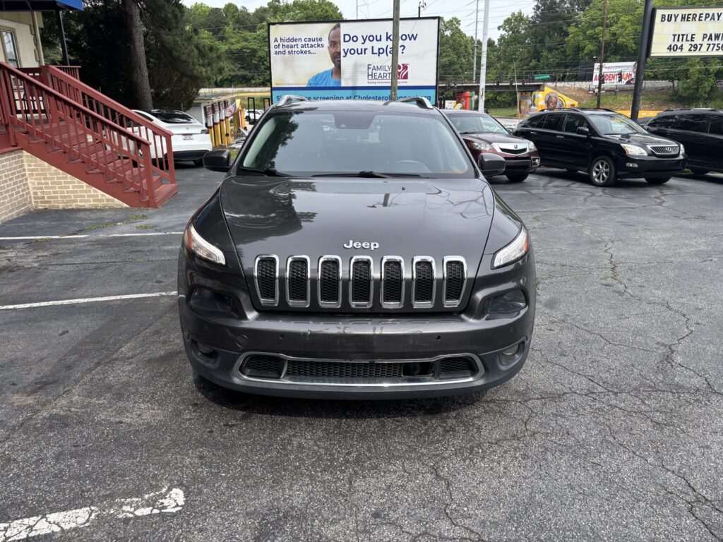 Front view of a dark gray Jeep SUV parked in a lot, with a large billboard behind and other cars nearby.