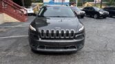 Front view of a dark gray Jeep SUV parked in a lot, with a large billboard behind and other cars nearby.