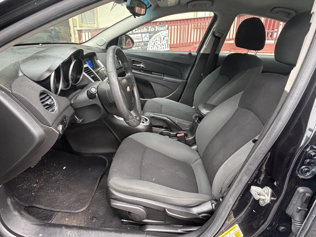 Interior of a black car showing driver's seat, steering wheel, and front passenger seat viewed from open driver-side door; dusty floor mats visible outside window at a drive-through area.