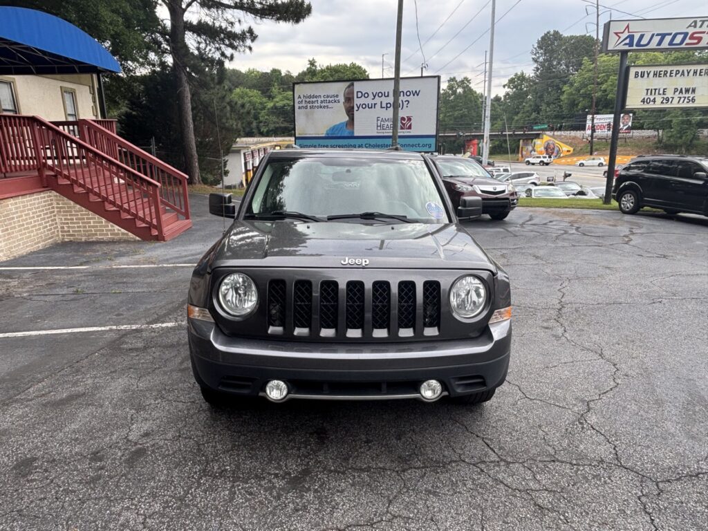 Front view of a gray Jeep SUV parked in a lot, with a blue building awning and red stairs on the left and billboards in the background.