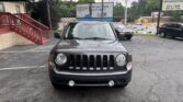 Front view of a gray Jeep SUV parked in a lot, with a blue building awning and red stairs on the left and billboards in the background.